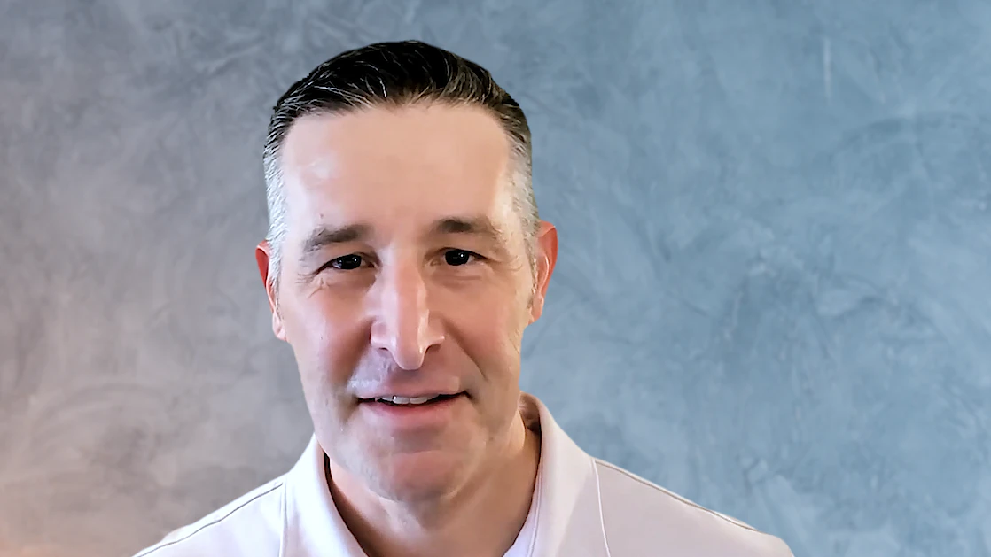 Professional man smiling in a light-colored shirt against a textured blue background, representing the human element of sales training and coaching at RAMPT Consulting.